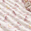 A close-up of a plush blanket or fabric featuring a repeating unicorn and cloud print on a warm off-white background. The design includes pastel pink and lavender unicorns, soft pink clouds, small gold stars, and gold crescent moons.