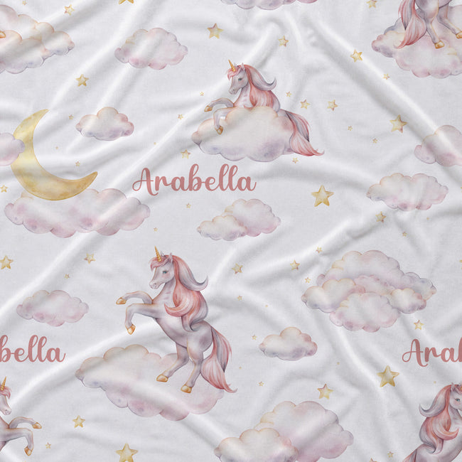White fabric with a whimsical unicorn print, featuring pastel pink and lavender unicorns on fluffy clouds, a gold crescent moon, small gold stars, and the personalized name 'Arabella' in pink script.