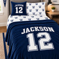 A navy blue and white varsity bedding set styled on a bed in a bedroom, featuring a large 'JACKSON 12' print on the comforter, matching pillowcases, and a separate decorative pillow, with a wooden nightstand and lamp in the background.