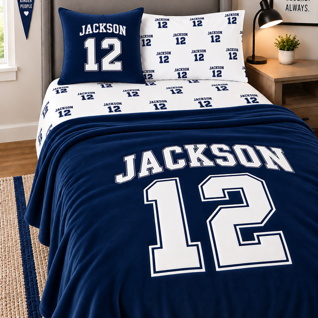 A navy blue and white varsity bedding set styled on a bed in a bedroom, featuring a large 'JACKSON 12' print on the comforter, matching pillowcases, and a separate decorative pillow, with a wooden nightstand and lamp in the background.