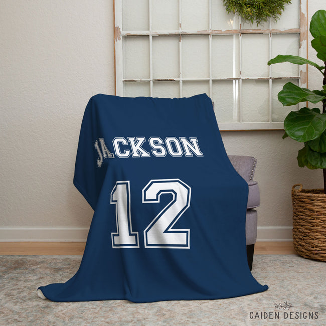 A navy blue throw blanket draped over a gray armchair in a living room setting, featuring the name 'JACKSON' and number '12' printed in white.
