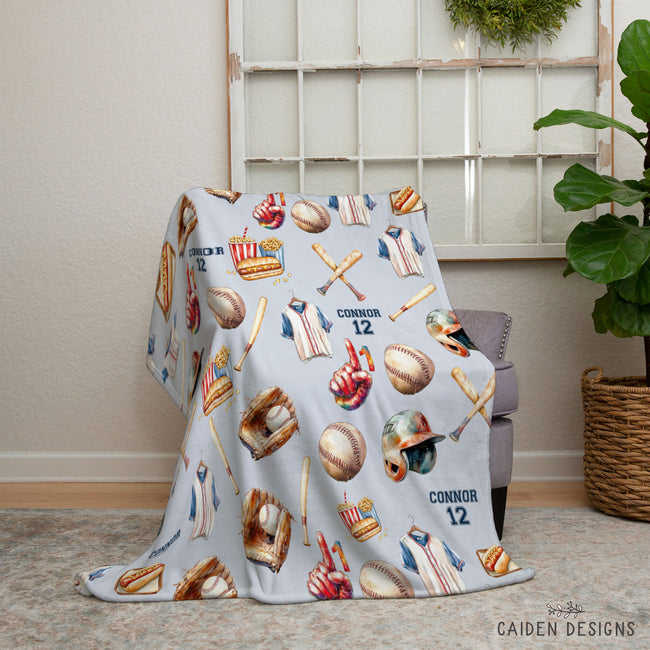 A light blue personalized baseball-themed throw blanket draped over a chair in a styled room setting. The blanket features a pattern of baseball jerseys, baseballs, bats, helmets, and glove details, with the names 'CONNOR 12' printed on it.