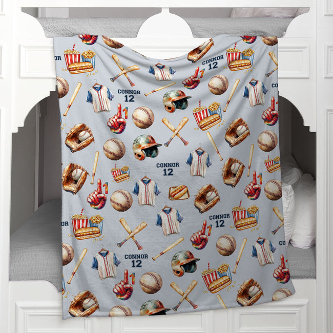 A light gray blanket featuring a baseball-themed pattern with baseballs, bats, jerseys, helmets, and the name 'CONNOR 12', draped over a white wooden bed frame.