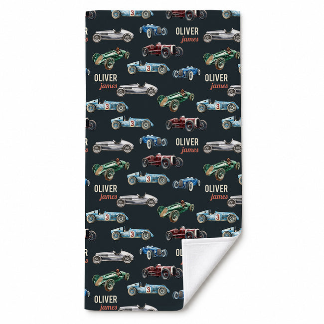 A beach towel with a dark navy background printed with vintage race cars and the personalized name 'OLIVER james' in a repeating pattern. The bottom right corner is folded over to show a white reverse side, set against a plain white background.