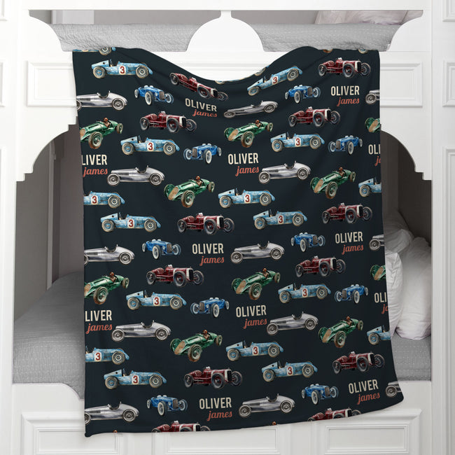 A personalized fleece blanket featuring a vintage race car pattern with the name 'OLIVER James' repeated, draped over a white wooden mantel.