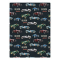 A blanket featuring a repeating pattern of vintage racing cars in red, blue, green, and silver on a dark charcoal background, with the personalized name 'Oliver James' printed in white and orange text.