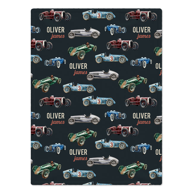 A blanket featuring a repeating pattern of vintage racing cars in red, blue, green, and silver on a dark charcoal background, with the personalized name 'Oliver James' printed in white and orange text.