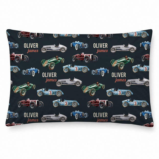 A pillowcase featuring a repeating pattern of vintage race cars in blue, green, red, and silver on a dark navy background, with the personalized text 'OLIVER james' printed in white and orange.