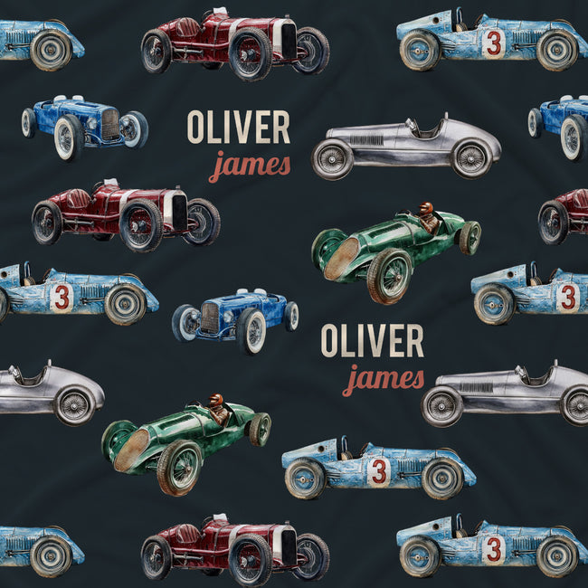 A dark navy fabric featuring a repeating pattern of vintage racing cars in blue, green, red, and silver, interspersed with the personalized text 'OLIVER james' in cream and coral.