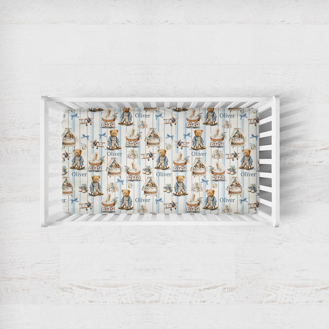 A baby crib with white slats, fitted with a printed sheet featuring teddy bears, boats, and the name 'Oliver' in a vintage illustration style, set against a light whitewashed wood background.