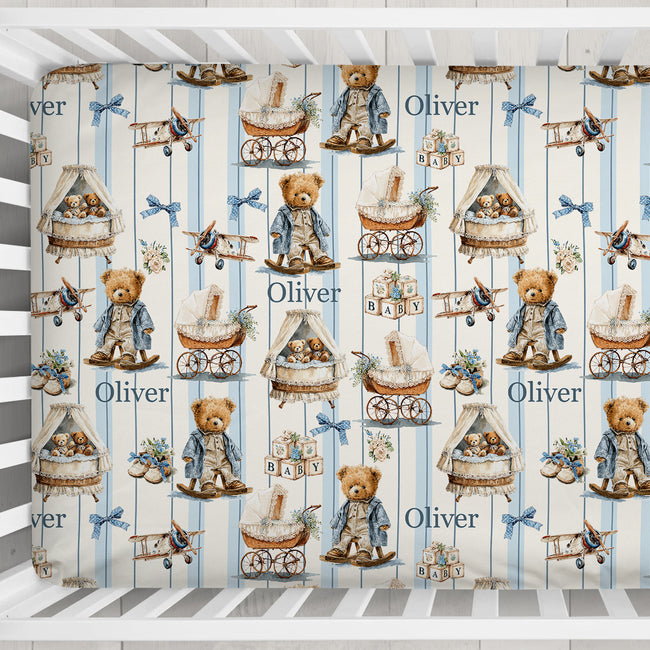 A crib bedding set featuring a repeating pattern of teddy bears, baby carriages, and the name 'Oliver' on blue and white vertical stripes, shown inside a white wooden crib.