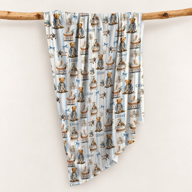 Personalized swaddle blanket hanging on a wooden rail with vintage teddy bears, baby carriages, toys and custom name Oliver.