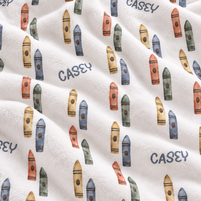 A close-up of a white plush fabric with an all-over print of colorful crayons and the name CASEY repeated in dark blue text.