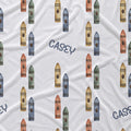 White fabric with a repeating pattern of colorful crayons in blue, green, orange, and yellow, interspersed with the name 'CASEY' in navy blue script.