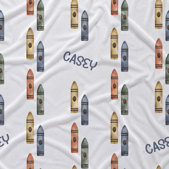 White fabric with a repeating pattern of colorful crayons in blue, green, orange, and yellow, interspersed with the name 'CASEY' in navy blue script.