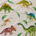 A close-up of a fabric surface printed with multiple illustrated dinosaurs in green, yellow, blue, brown, and pink, plus scattered green leaves on an off-white background.