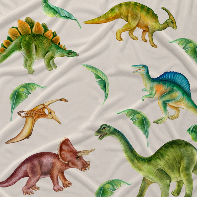 A close-up of a fabric surface printed with multiple illustrated dinosaurs in green, yellow, blue, brown, and pink, plus scattered green leaves on an off-white background.