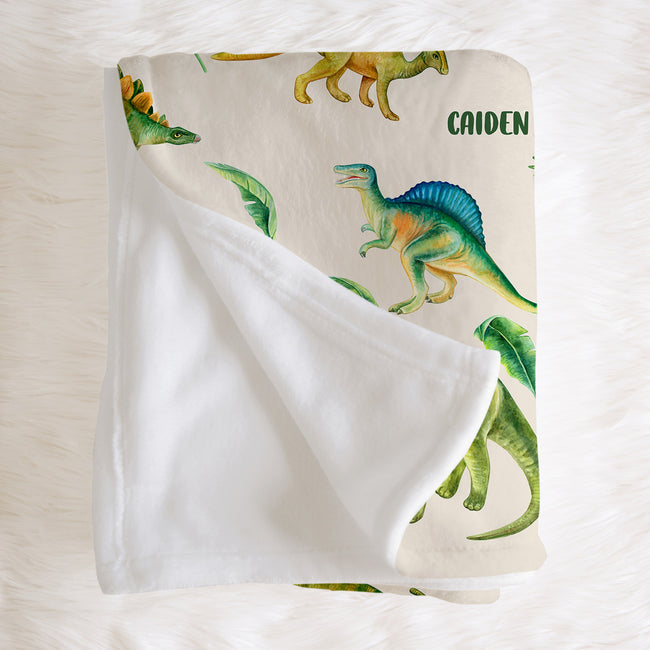 A personalized baby blanket with a white sherpa backing and a printed dinosaur pattern on a cream background, laid flat on a white faux fur surface. The name 'CAIDEN' is printed in green near the top right.