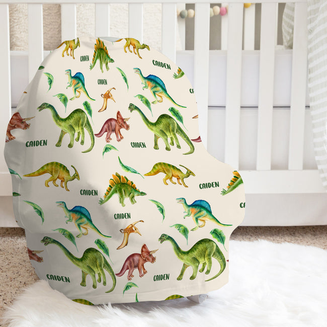 A baby car seat cover featuring a colorful print of various dinosaurs on an off-white background with the name 'Caiden' repeated, styled in a nursery setting in front of a white crib.