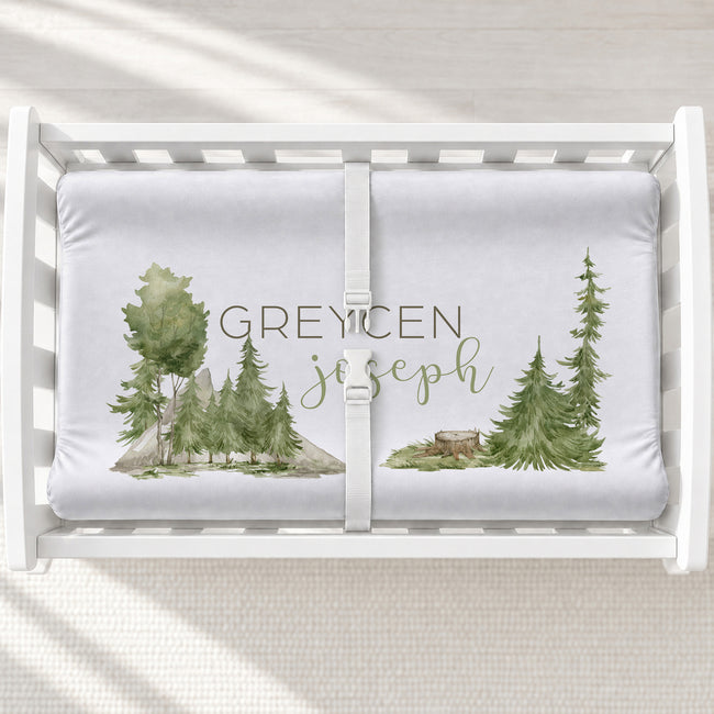 A white changing pad printed with a watercolor forest design and the personalized name 'GREYCEN joseph' in muted green and brown lettering.