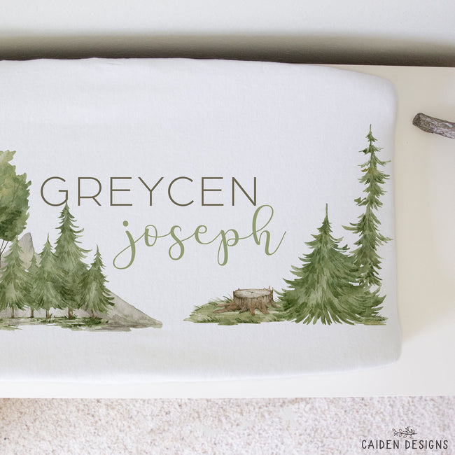 A white changing pad printed with a watercolor forest design and the personalized name 'GREYCEN joseph' in muted green and brown lettering.