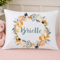 A personalized white pillowcase featuring a watercolor floral wreath and the name 'Brielle' in teal script, styled on a pink quilted bed next to a pink teddy bear.
