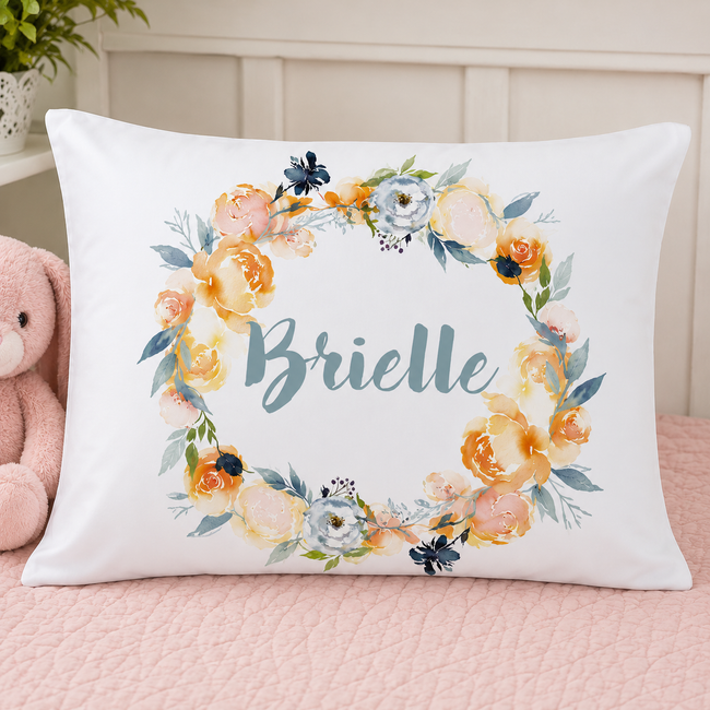 A personalized white pillowcase featuring a watercolor floral wreath and the name 'Brielle' in teal script, styled on a pink quilted bed next to a pink teddy bear.