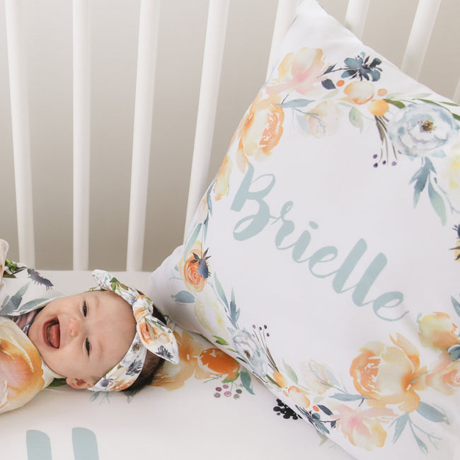 A baby lying on a white crib mattress wearing a matching floral headband, next to a large floral pillow printed with the name Brielle in script.