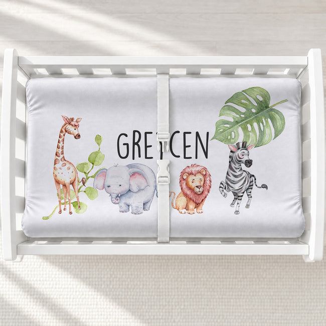 A personalized white changing pad featuring a watercolor giraffe (off-screen), elephant, lion, zebra and plants with the name "GREYCEN" in black, capitalized letters.