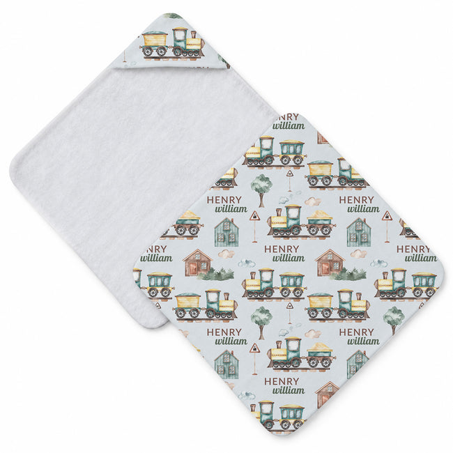 A personalized baby hooded towel with a white background and a repeating pattern of vintage train illustrations in green, yellow, and brown, with the name 'HENRY' and 'william' printed throughout. The front panel is folded over to show the soft, textured light gray fabric backing.