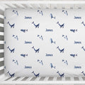 A white fitted crib sheet with a repeating pattern of blue whales and the name 'James' printed in blue, shown inside a white wooden crib.