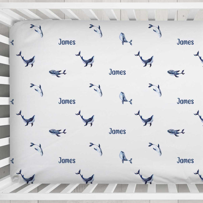 A white fitted crib sheet with a repeating pattern of blue whales and the name 'James' printed in blue, shown inside a white wooden crib.