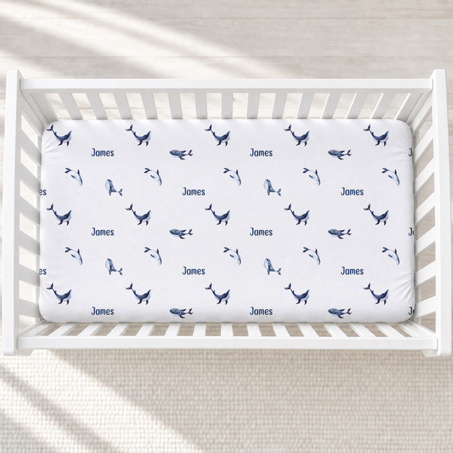 A white crib mattress fitted sheet with a navy blue whale print and the name 'James' repeated, shown inside a white wooden crib on a light neutral floor.