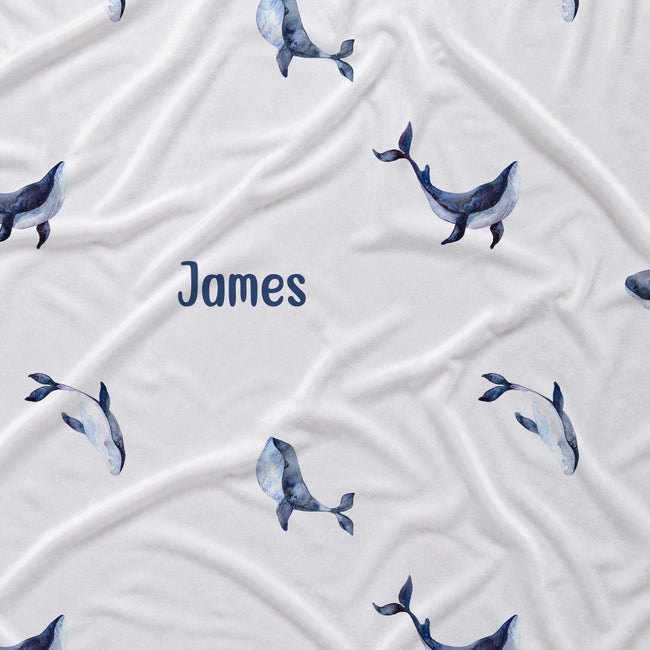 A white fabric item, likely a baby blanket or swaddle, featuring a repeating pattern of watercolor blue and gray whales and the name 'James' printed in navy blue text in the center.