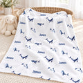 A white personalized blanket with blue whale prints and the name 'James' repeated, draped over a woven basket in a bright nursery setting.