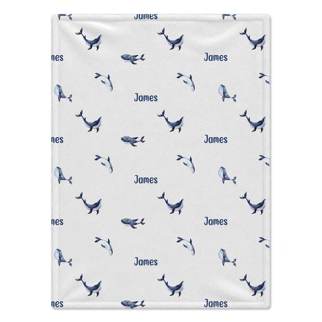A personalized baby blanket with a white background, featuring a repeating pattern of blue whales and dolphins, along with the name 'James' printed in blue. The blanket is shown flat against a clean white background.