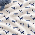 A close-up of a plush white blanket with a repeating pattern of blue whales and the name 'James' printed in blue, with a gray whale toy partially visible in the top-left corner.