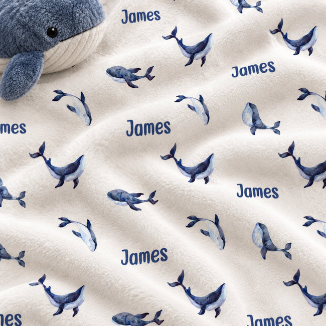 A close-up of a plush white blanket with a repeating pattern of blue whales and the name 'James' printed in blue, with a gray whale toy partially visible in the top-left corner.