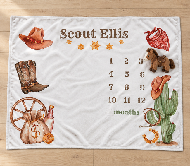 A personalized fleece blanket with a western theme, laid flat on a light wood floor. The blanket features the name 'Scout Ellis' at the top, a 12-month countdown grid, and various western illustrations including a cowboy hat, boots, cactus, and money bag.