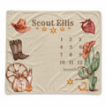 A personalized cream-colored blanket featuring a western-themed design with the name 'Scout Ellis', a 12-month calendar grid, and illustrations of a cowboy hat, boots, cactus, and other cowboy motifs.