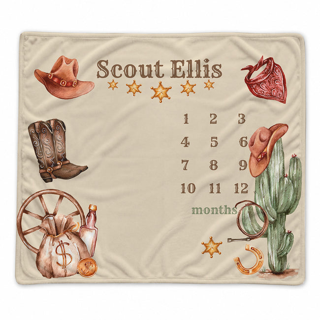 A personalized cream-colored blanket featuring a western-themed design with the name 'Scout Ellis', a 12-month calendar grid, and illustrations of a cowboy hat, boots, cactus, and other cowboy motifs.