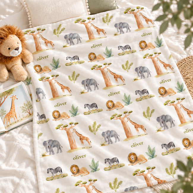 Wildwood Safari blanket personalized with Oliver, spread on white bedding beside a plush lion and an open giraffe picture book.