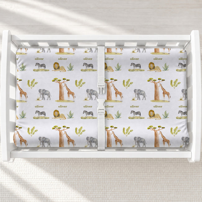 Wildwood Safari changing pad cover with watercolor elephants, giraffes, lions, zebras, and the name Oliver, shown on a white changing table.