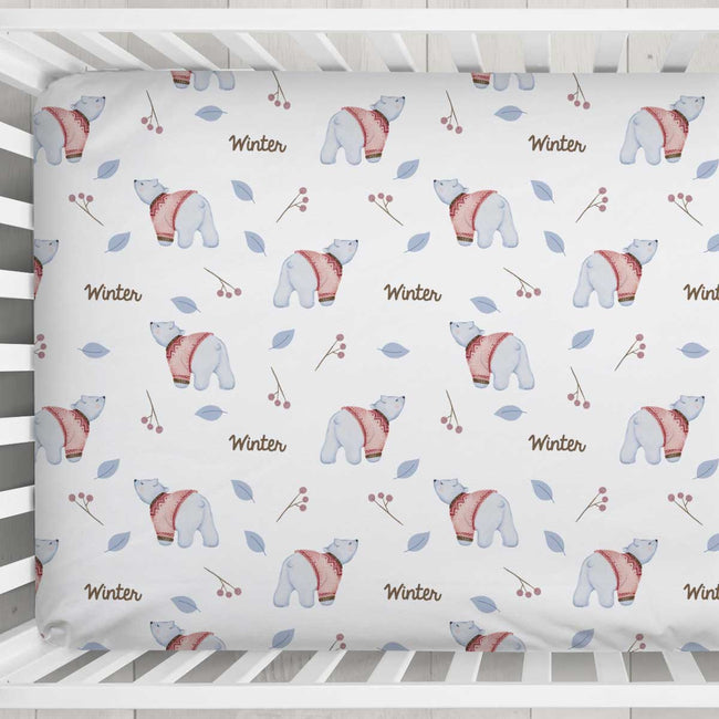 A fitted crib bumper or mattress cover with a winter-themed pattern of polar bears, berries, leaves, and the word 'Winter' in brown script, shown inside a white wooden crib.