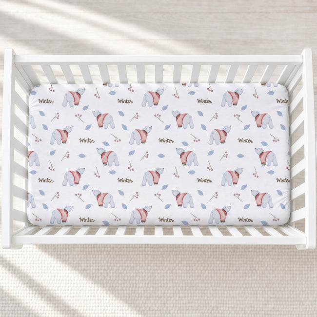 A fitted crib mattress cover with a winter polar bear pattern, shown inside a white wooden crib on a light textured blanket.