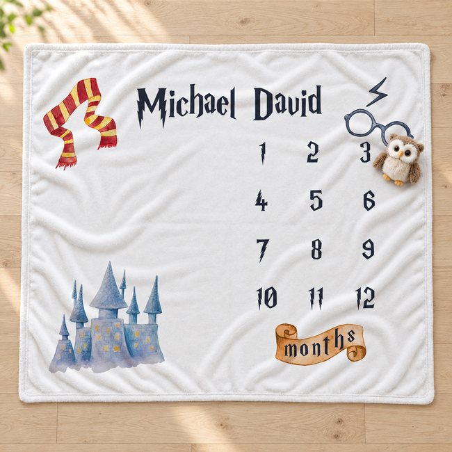 A personalized baby blanket laid flat on a light wood floor. The white blanket features a wizard theme with a red and yellow striped scarf, a blue castle, black text reading 'Michael David' and numbered days of the week, a small owl plush, and a gold banner that says 'months'.