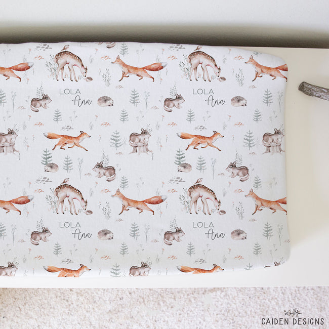 A personalized changing pad cover featuring a woodland print with foxes, deer, squirrels, hedgehogs and pine trees on a white background. The text 'LOLA Ann' is printed in a script font.