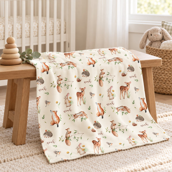 A cream-colored baby blanket with a woodland animal print featuring foxes, deer, hedgehogs, and acorns, draped over a wooden bench in a nursery setting.