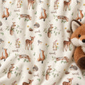 A close-up of a cream-colored fabric with a repeating woodland print featuring brown foxes, deer, rabbits, squirrels, hedgehogs, birds, and the name 'noah' in script. A plush brown fox toy is partially visible on the right side.