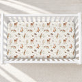 A fitted crib sheet with a woodland animal print is shown inside a white wooden crib, viewed from above. The print features brown deer, foxes, rabbits, hedgehogs, and squirrels with green leaves on an off-white background.